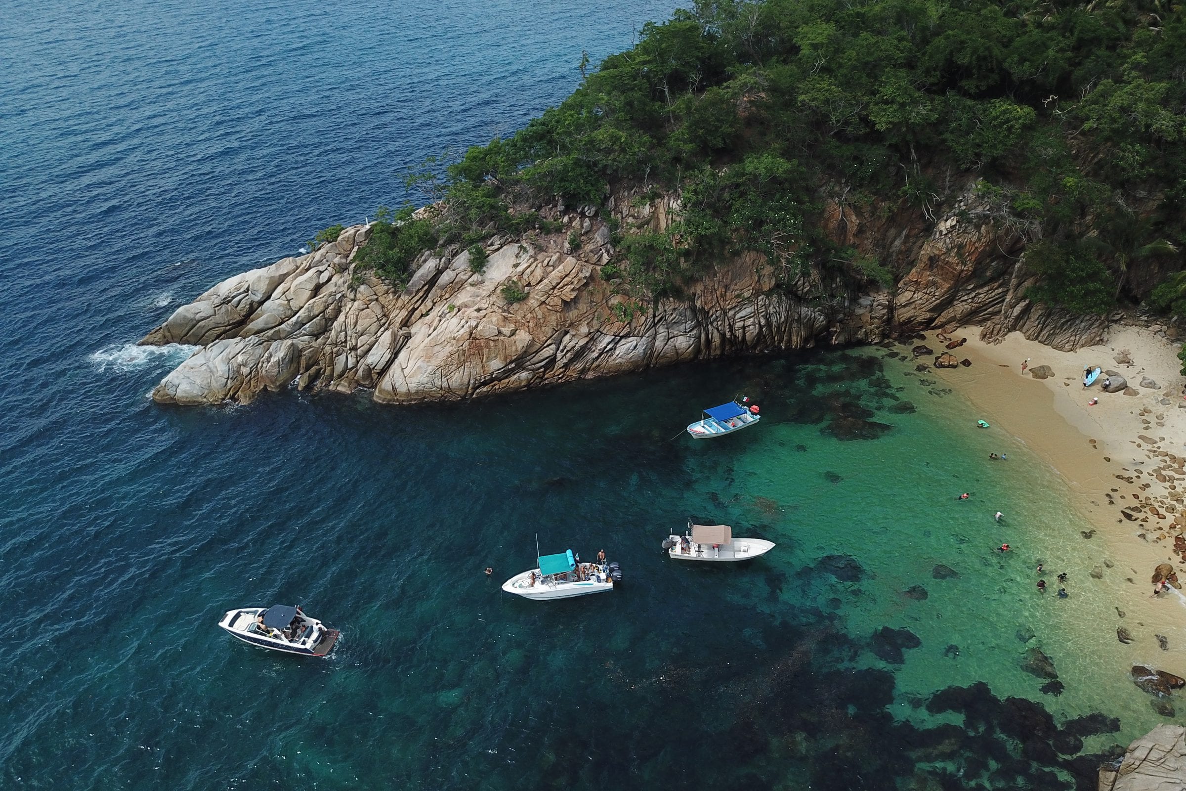Vista aérea de una cala de Bahía de Banderas: punta rocosa con selva, agua turquesa, playa y lanchas fondeadas.
